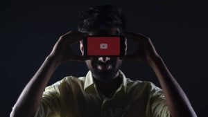a person is holding a phone against his eyes, the screen is looking toward the camera taking the image and it is displaying the youtube starting screen, the logo, it is taken in a black room, with low lighthing