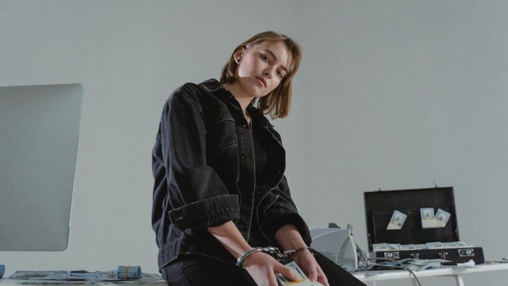 delito de alzamiento de bienes, una chica esta sentada en una mesa con muchos billetes, tambien hay un matein y esta esposada, refleja una situacion donde "te han pillado"