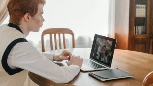 Red-haired boy doing a video conference with a laptop on a living room table, next to it is a notepad.