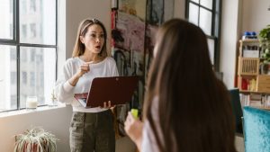 una mujer le esta explicado algo a una chica en el salon de un piso, la mujer tiene un portatil en las manos y brazos y esta de pie