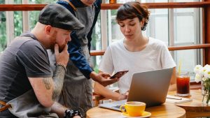 como optimizar google my business, tres personas interactuando con un movil y un portatil en una cafeteria moderna y estilosa, con mesas y ventanas de madera
