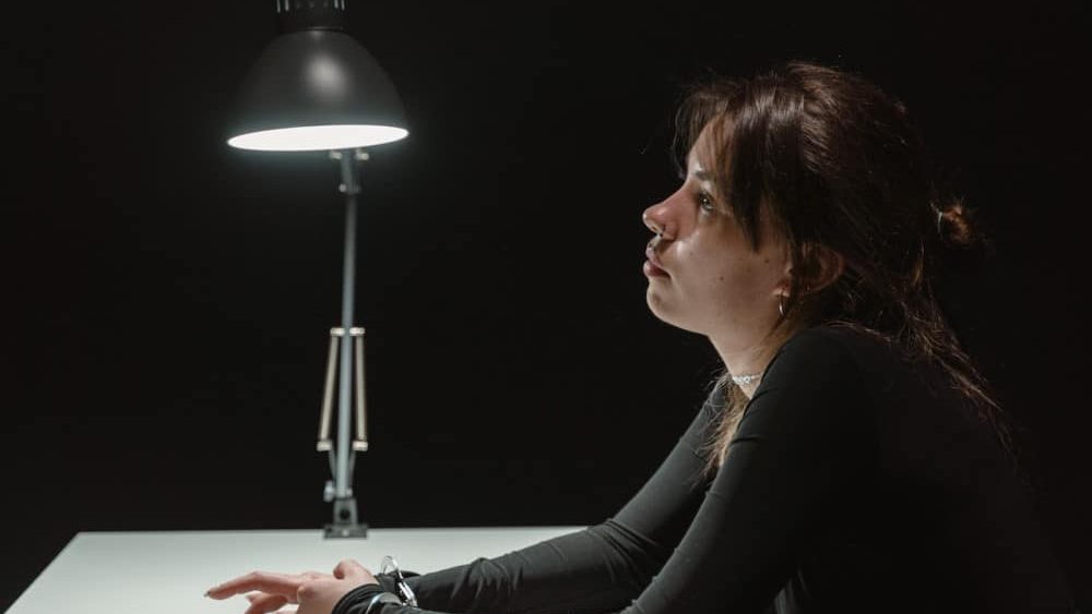 que es habeas corpus, una chica esta sentada en una silla enfrente de una mesa blanca esposada con una lampara a la derecha, el fondo es oscuro