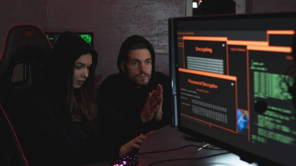 que es un malware, dos personas estan sentadas frente a ordenadores, el ambiente es oscuros, y bastante "cyber", parece una habitacion de un hacker