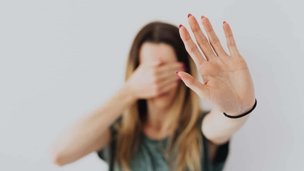una chica alza la mano erguida hacia delante y con la otra se tapa la cara el fondo es muy claro