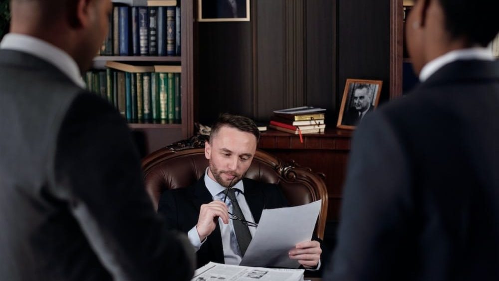 que es un term sheet, 3 abogados o hombres de negocio estan en una reunion infromal, pues 2 estan de pie y el otro esta sentado, mirando un docuemtno y con la patilla de las gafas en la boca, estil pensativo