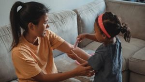 una niña chica le pinta con una jeringuilla de juguete a otra chica mas mayor en un sofa de una habitacion