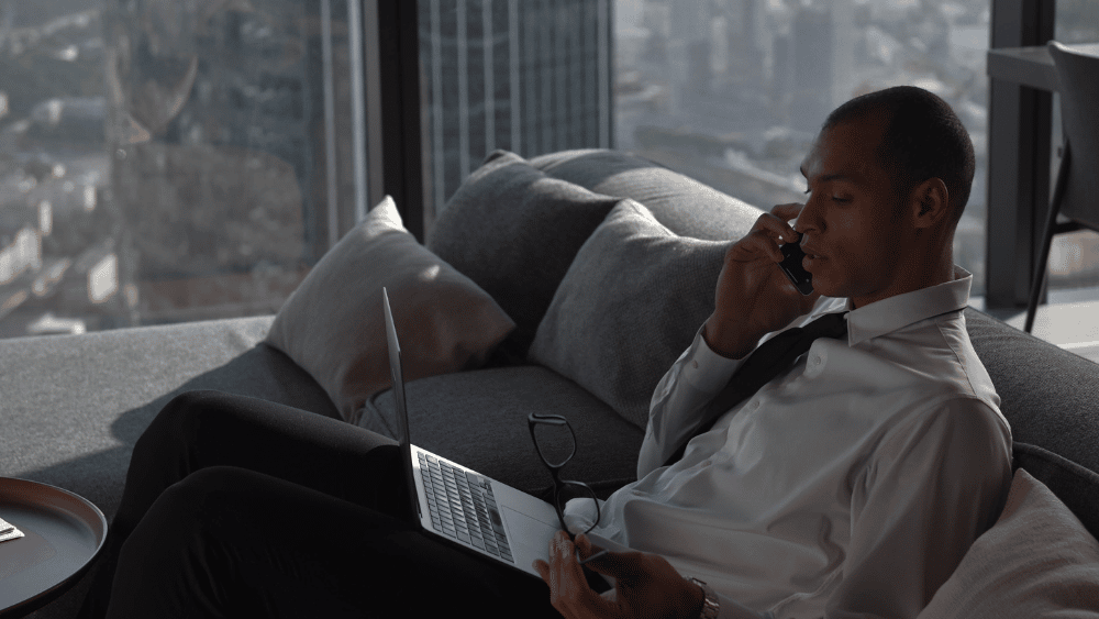 persona habla por el movil sentado en un sofa con vistas a la ciudad que se ven en el ventanal de atras, tambien tiene un portatil en las piernas
