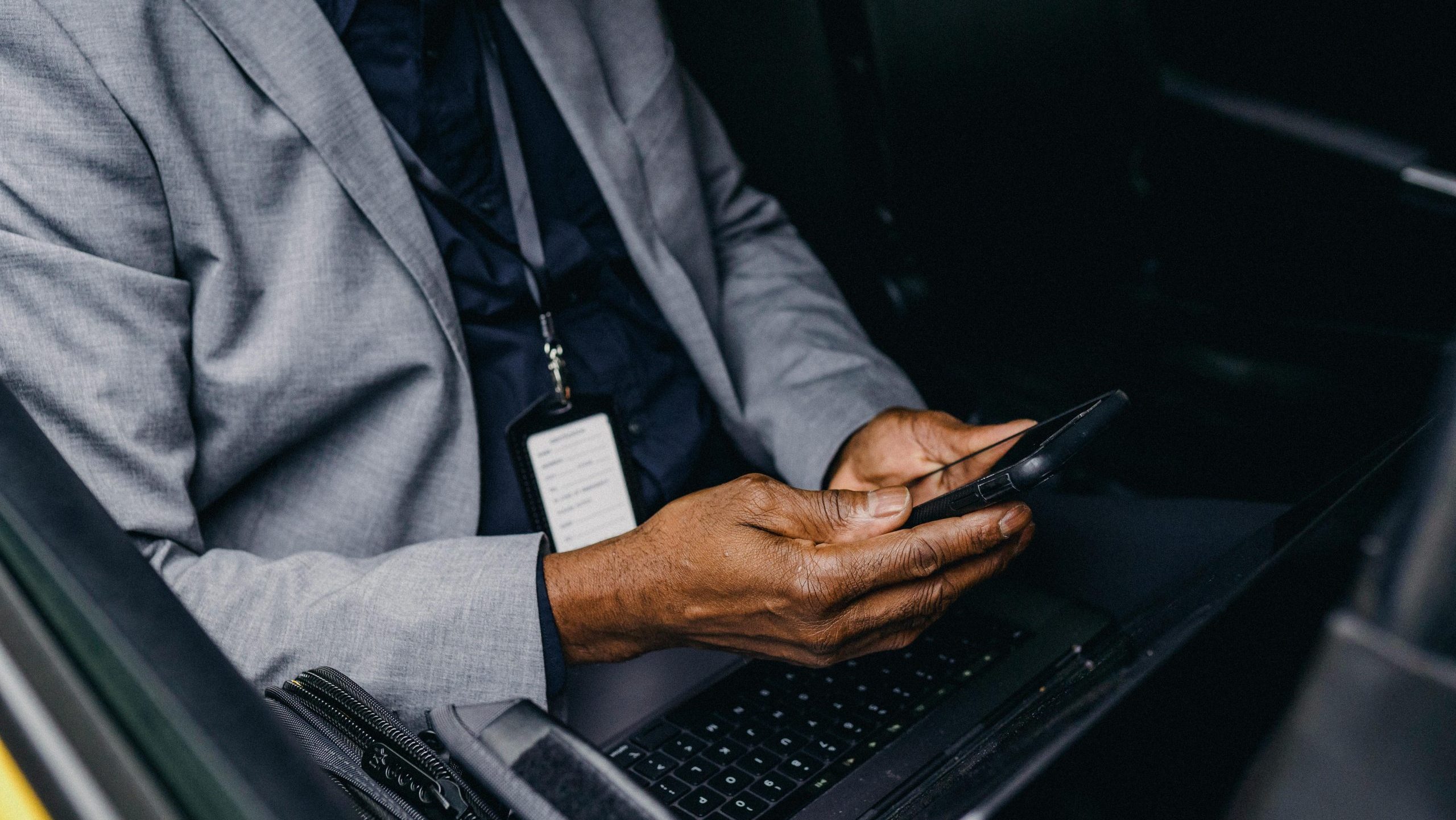 A person sitting in a taxi with a laptop on their lap and holding a mobile phone. They are wearing a suit, and a white badge with information hangs around their neck like a necklace, although the text on the badge appears blurry due to the image having a bokeh effect.
