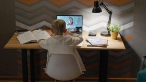 un chico pequeño estudia en una mesa de madera drente a un portatil con la camara encendida, esta haciendo una videollamada con una profesora