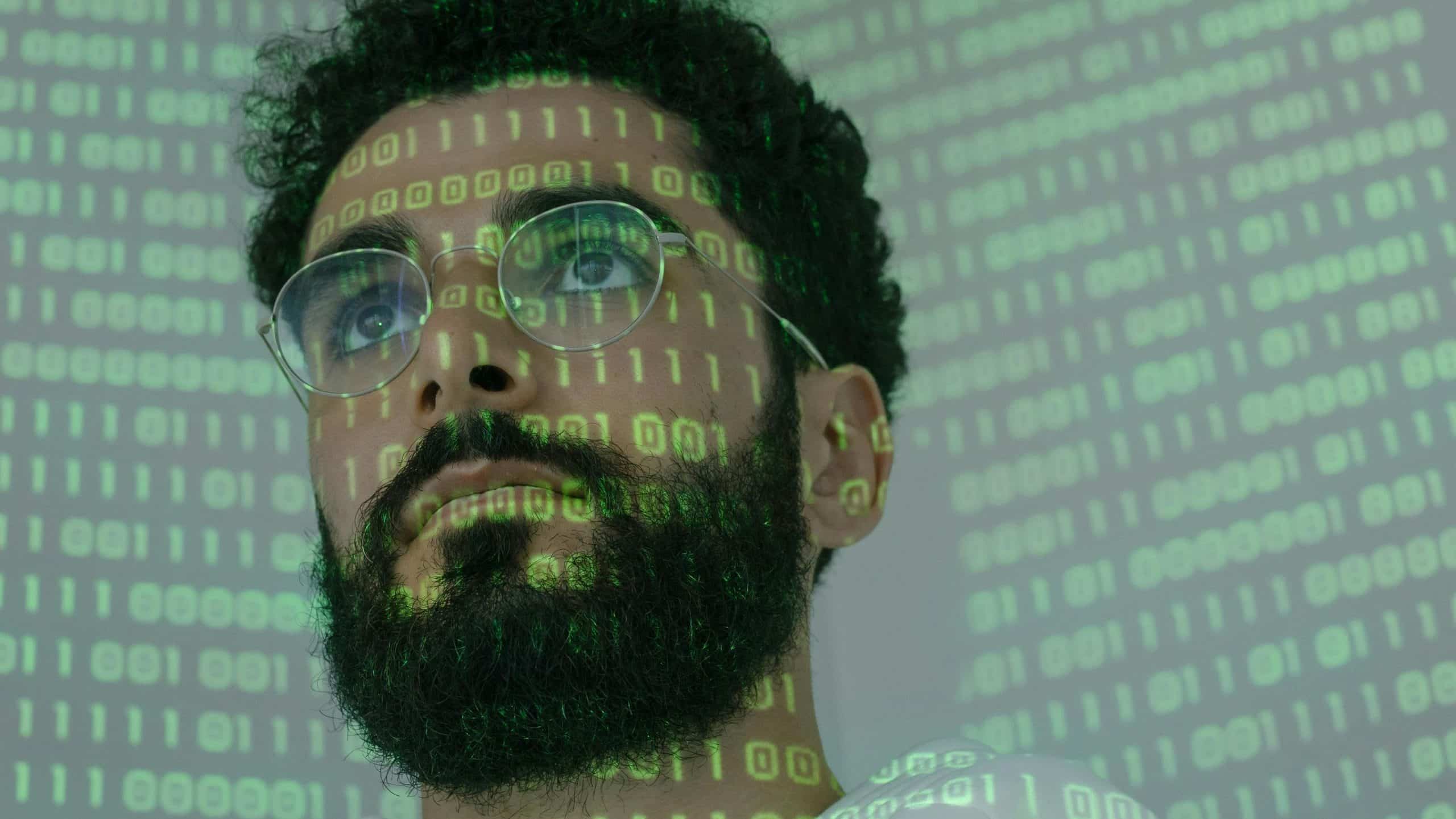 chico con gafas y barba, tiene numeros binarios siendo proyectados en su rostro y en la pared de atras