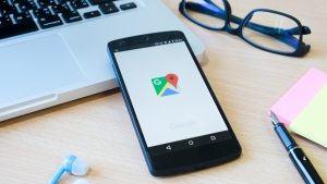 el inicio de google maps se ve en un movil apoyado contra un portatil en una mesa de madera junto con unas gafas un boli, postits, y unos auriculares
