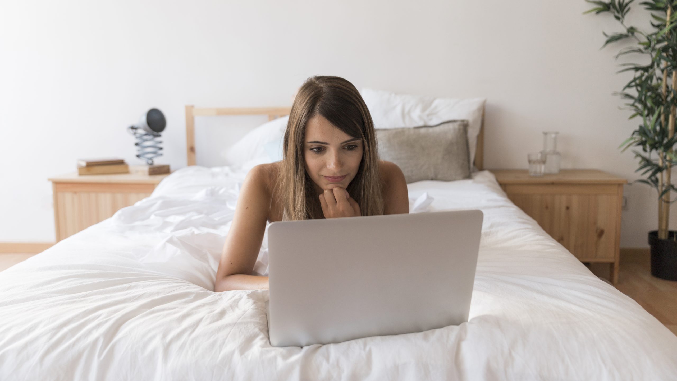 una chica esta tumbada boca abajo ante un portatil en la cama, el ambiente es calido. Esta en un cuarto