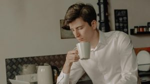 There is a boy in a kitchen holding a cup and looking down at something that is outside the image.