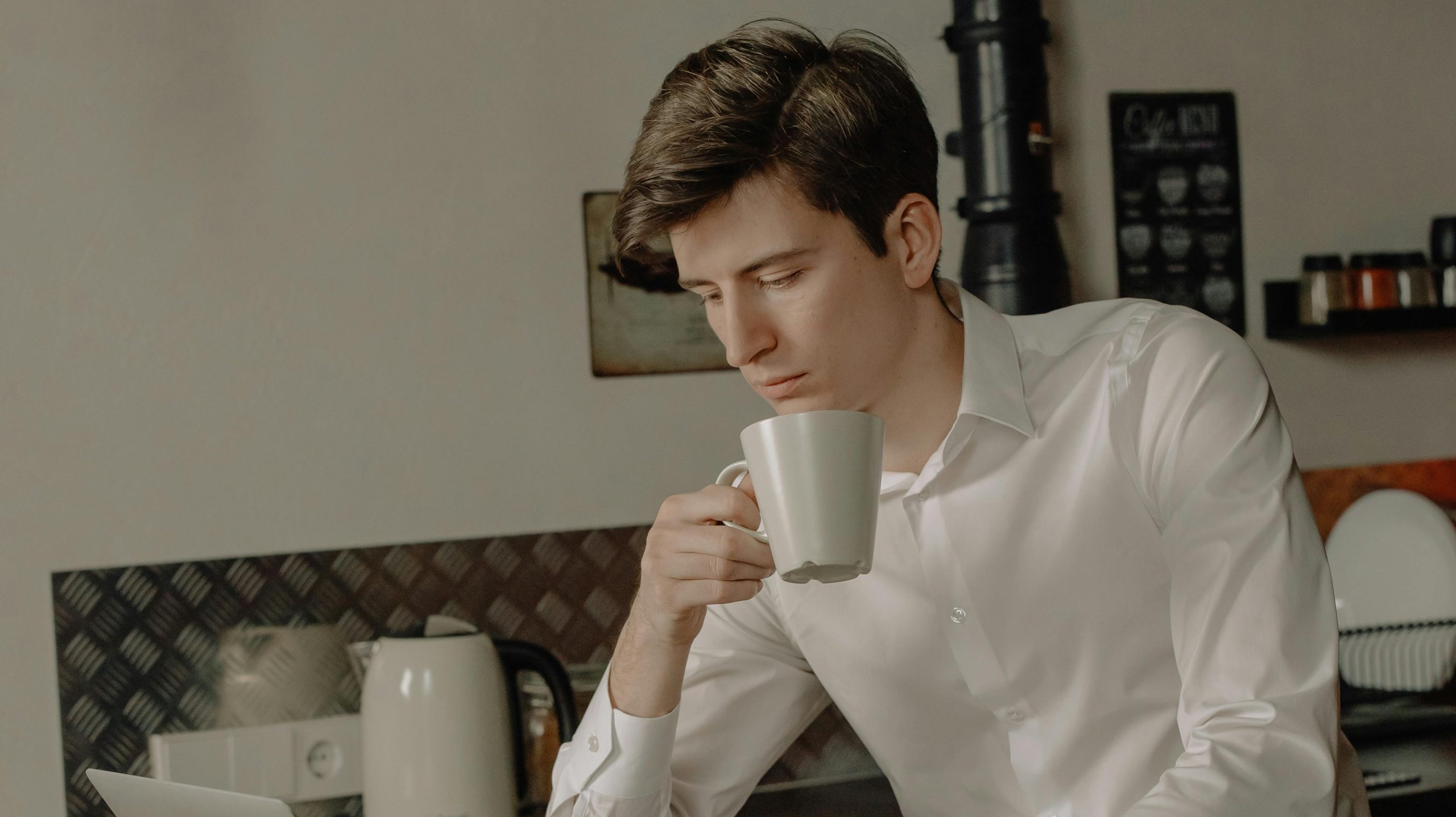 There is a boy in a kitchen holding a cup and looking down at something that is outside the image.