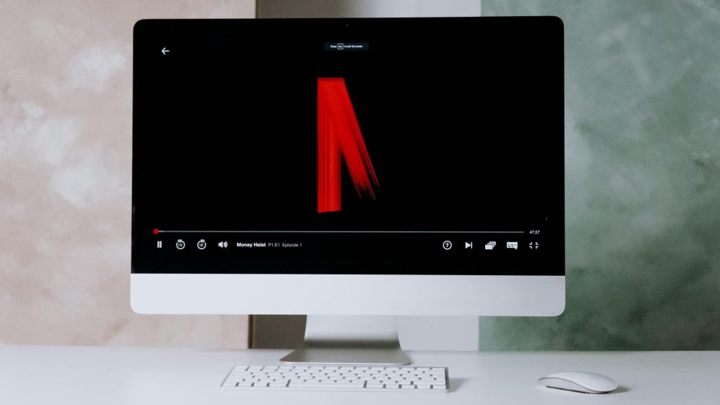 The background is white. The image is taken from an iMac with a keyboard and mouse. On the screen, the Netflix logo appears as a series begins.