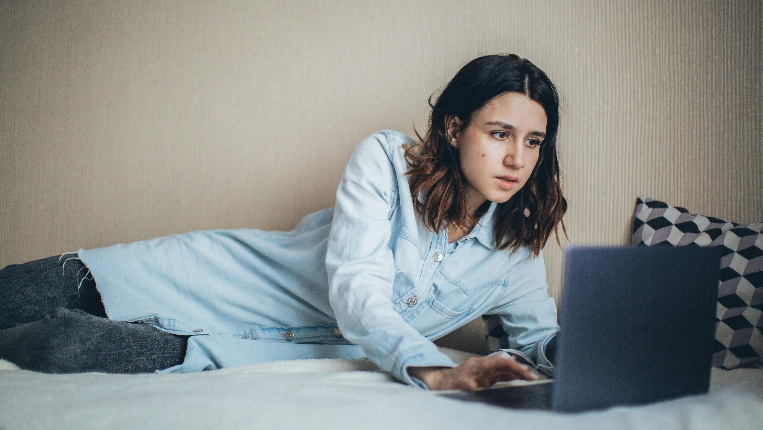 A girl lying on her side on a bed looks at a laptop with a suspicious expression. She's wearing a blue shirt and jeans.