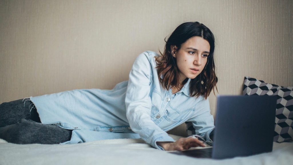 una chica tumbada de lado en una cama mira un portatil con rostro sospechoso, tiene una camisa azul y unos pantalones vaqueros