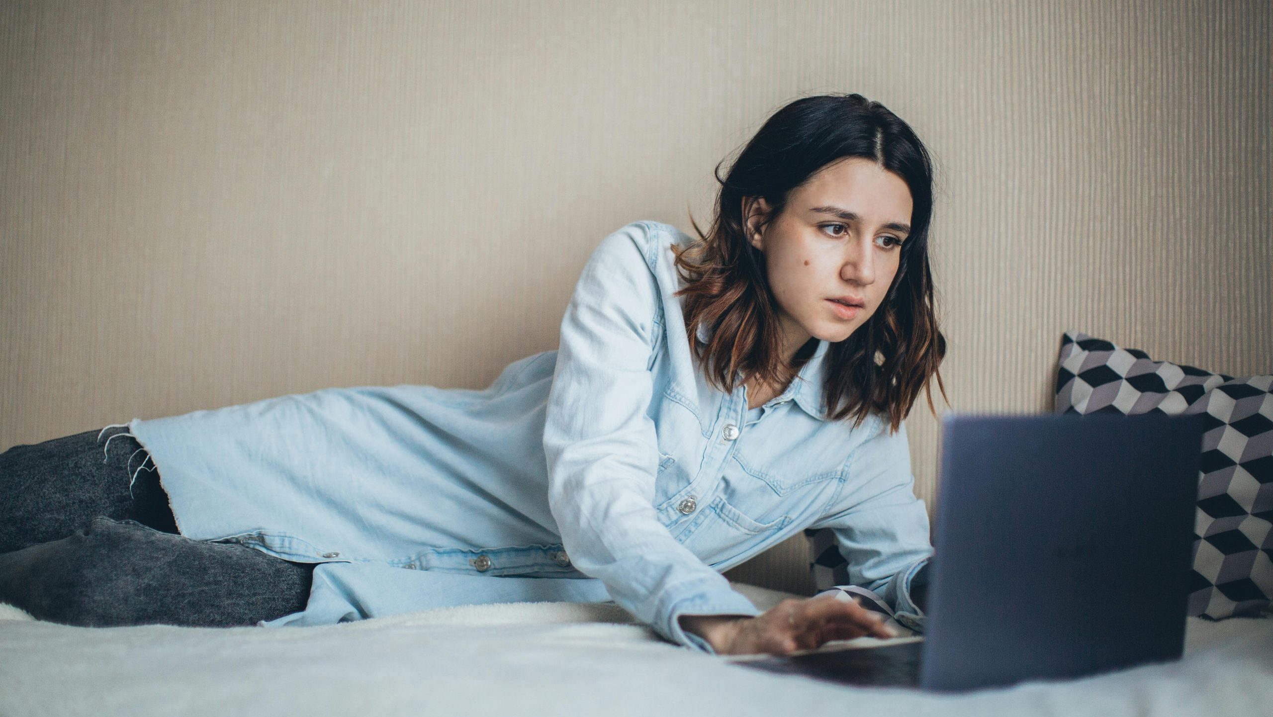 una chica tumbada de lado en una cama mira un portatil con rostro sospechoso, tiene una camisa azul y unos pantalones vaqueros