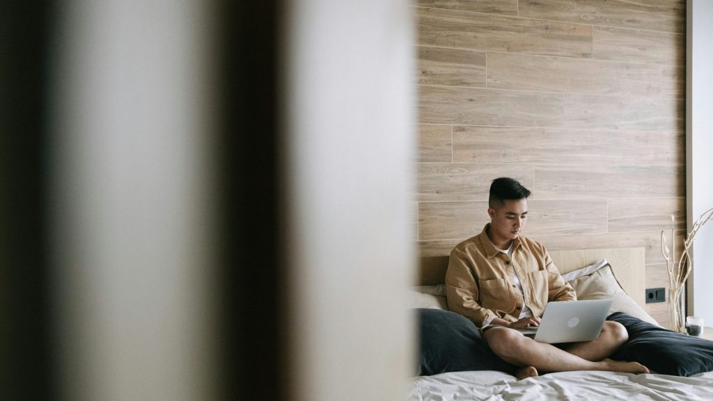 A person is using a laptop in bed. Sixty percent of the image is covered by a wall in the foreground, symbolizing that this person is in another room and that someone is observing. This wall is blurred or has bokeh. The focus is on him, who is sitting cross-legged on a bed. The photo is taken, and the room is lit. He has the laptop resting on his crossed legs.