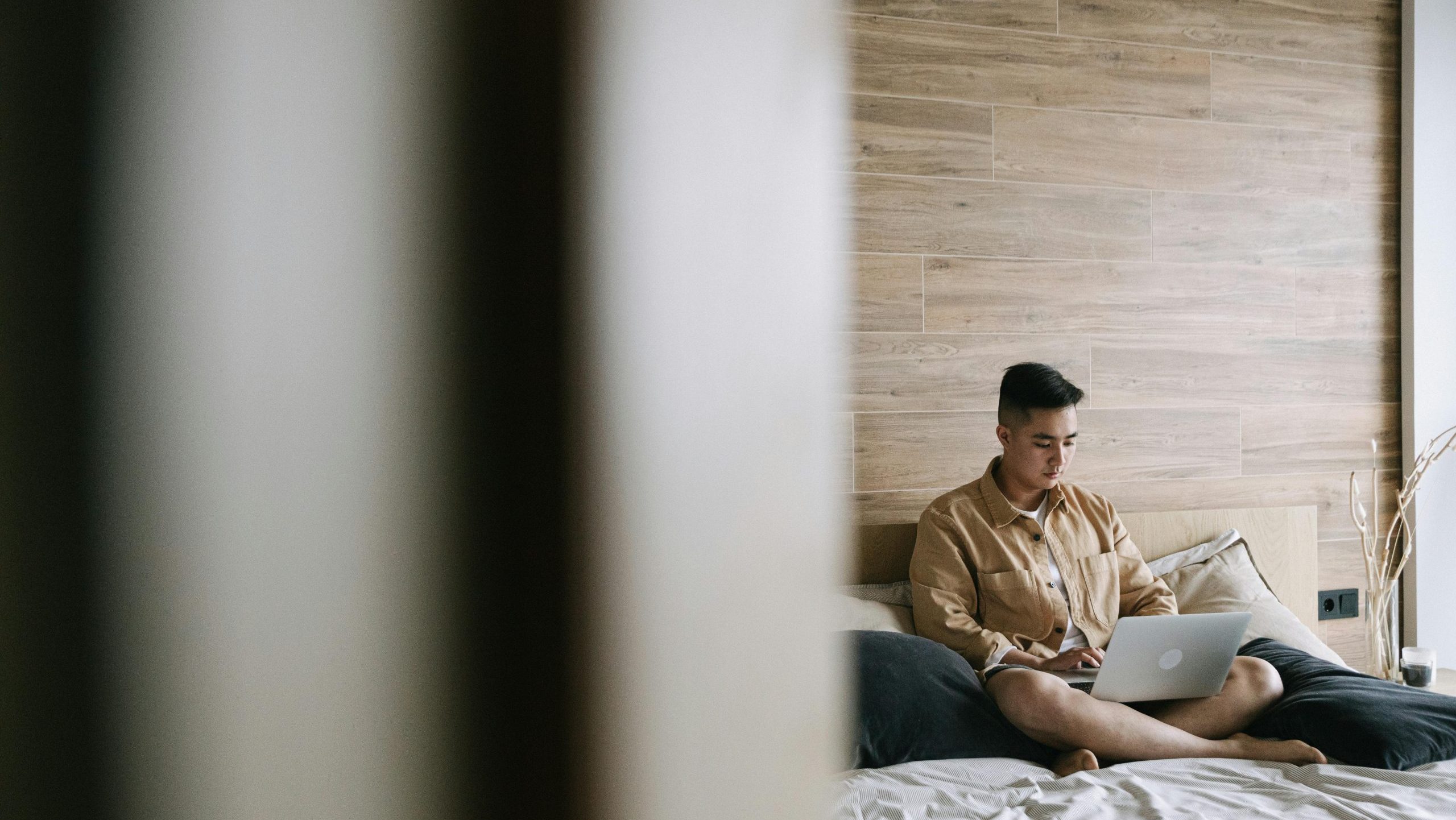 A person is using a laptop in bed. Sixty percent of the image is covered by a wall in the foreground, symbolizing that this person is in another room and that someone is observing. This wall is blurred or has bokeh. The focus is on him, who is sitting cross-legged on a bed. The photo is taken, and the room is lit. He has the laptop resting on his crossed legs.