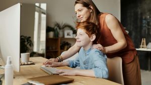 A woman is standing behind a boy, touching his shoulder with one hand. They are both looking at the computer screen. He is using the keyboard and mouse of the computer.