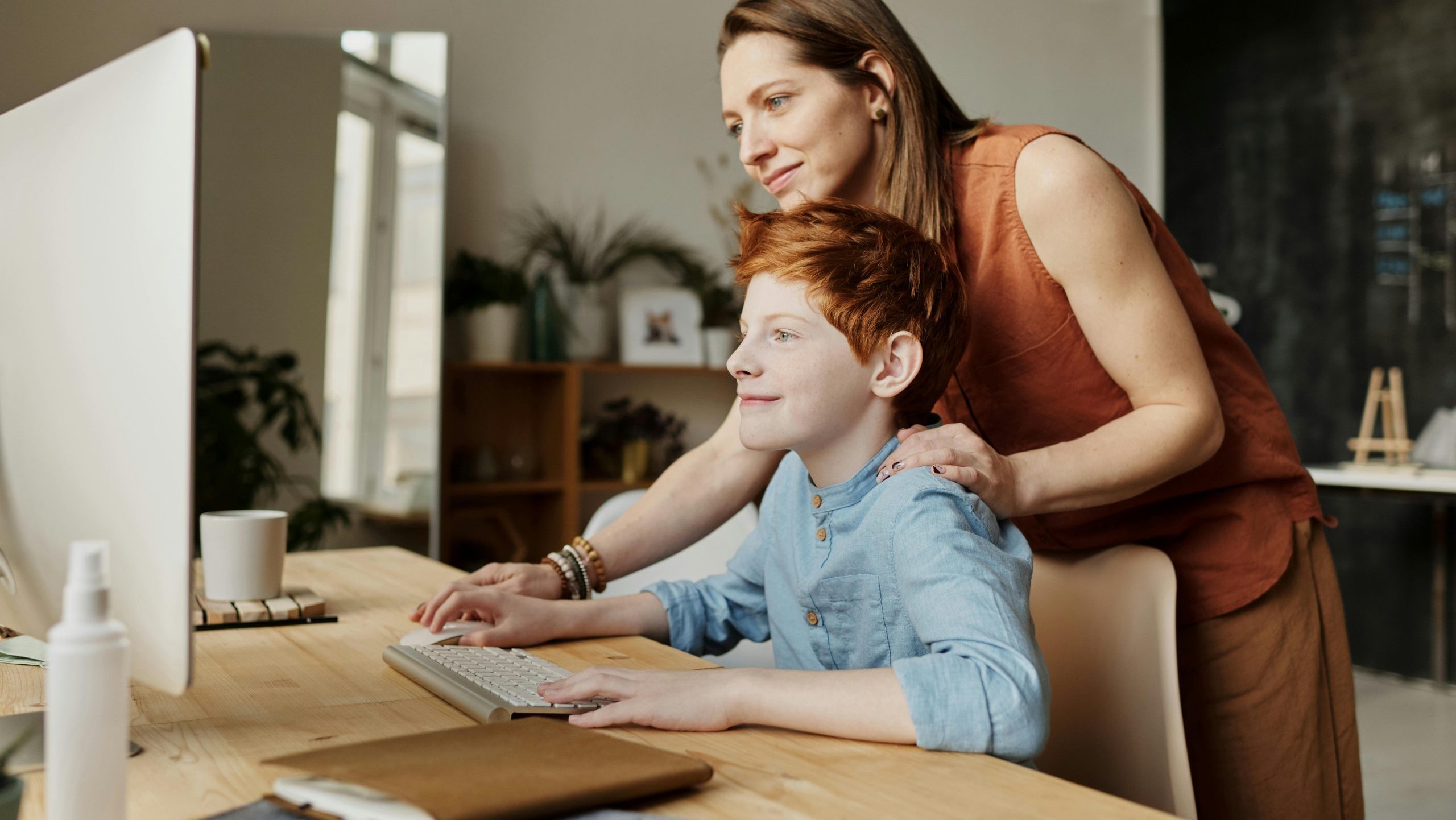 A woman is standing behind a boy, touching his shoulder with one hand. They are both looking at the computer screen. He is using the keyboard and mouse of the computer.