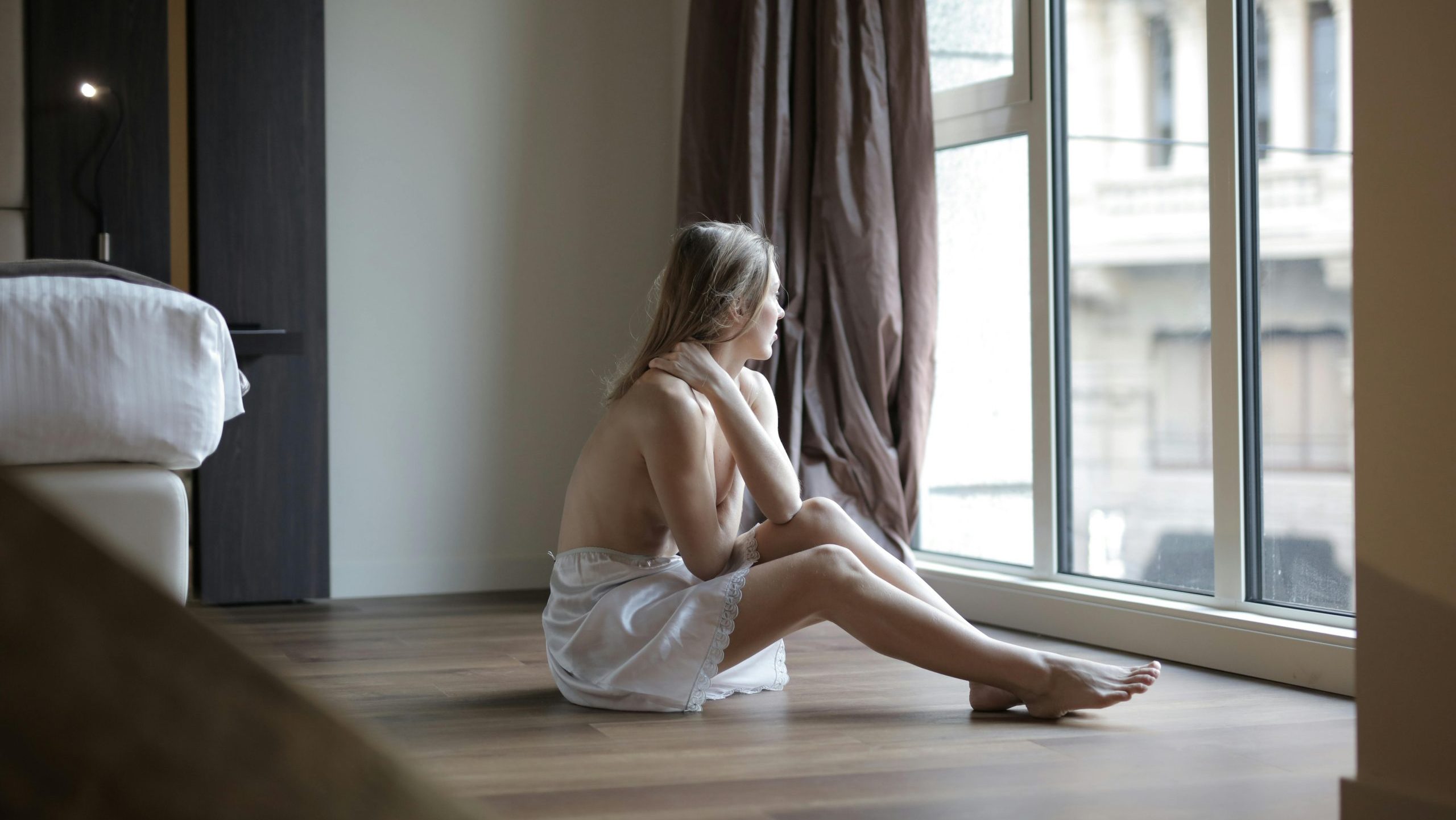 hay una chica sentada ene el suelo al lado de una cama semidesnuda ya que tiene el torso descubierto, esta en lo que podria ser una habitacion de un hotel y mira hacia afuera por la ventana