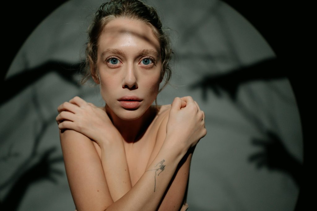 A girl with arms crossed in a way that covers her torso, she has a fixed gaze straight ahead with eyes wide open. Behind her, against a grayish wall, shadows of hands and arms can be seen playing with some threads.