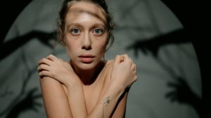 chica con brazos cruzados de manera que tapa su torso, tiene la mirada fija al frente con los ojos muy abiertos. Detras de ella ante una pared grisacea se ven las sobras de manos y brazos jugando con unos hilos