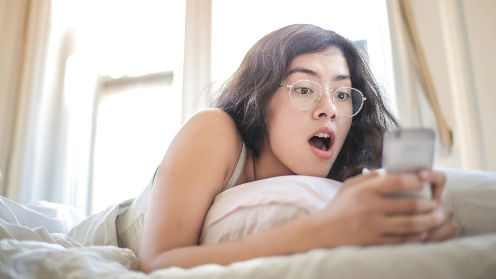 mujer usa el movil en la cama con cara de sorprendida, lleva gafas y el ambiente es cálido