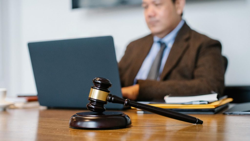 hay un hombre desenfocado en el fondo viendo un portatil, en primer plano hay un mazo legal
