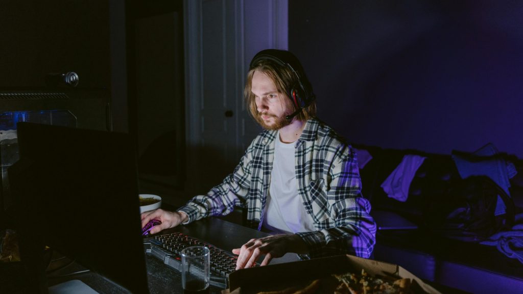 un hombre con el pelo largo tiene puestos unos cascos y usa el ordenador con el raton y el teclado, tanto el raton como el teclado estan iluminados, el lleva una camisa con una camiseta debajo y mira la pantalla, hay una caja de pizza a su izquierda con trozos de pizza dentro, la luz de la habitacion esta apagada, por eso el fondo es oscuro.