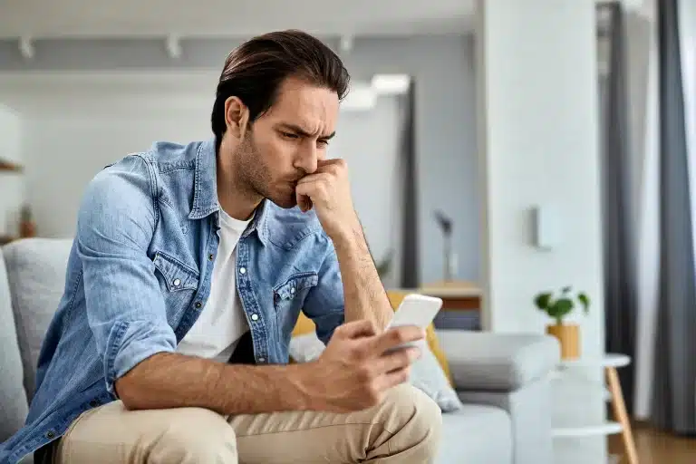 Chico preocupado en el sofá leyendo en el móvil un perfil falso que suplanta su identidad
